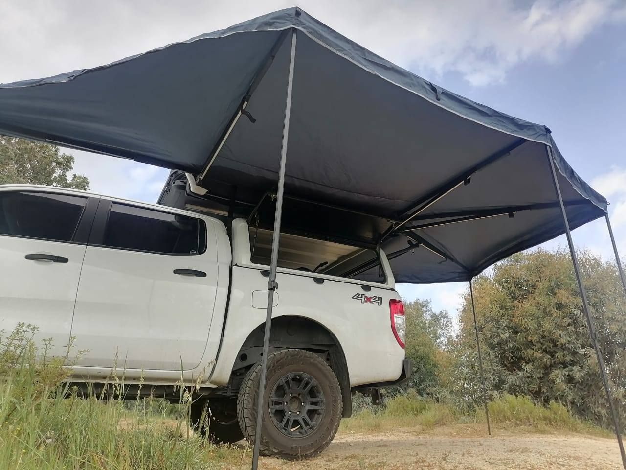 Batwing awning pitched on poles beside a white pickup on a grassy dirt trail