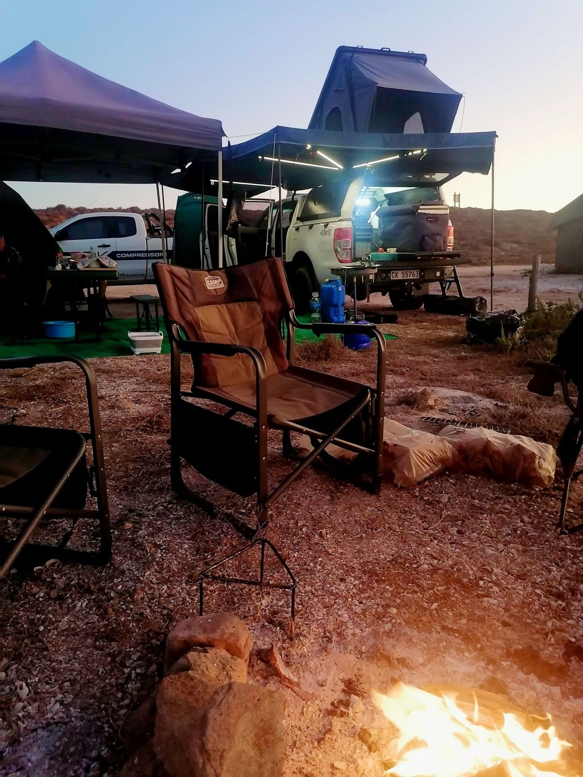 Evening camp — fire pit and camp chair facing a white pickup with lit awning and open tent