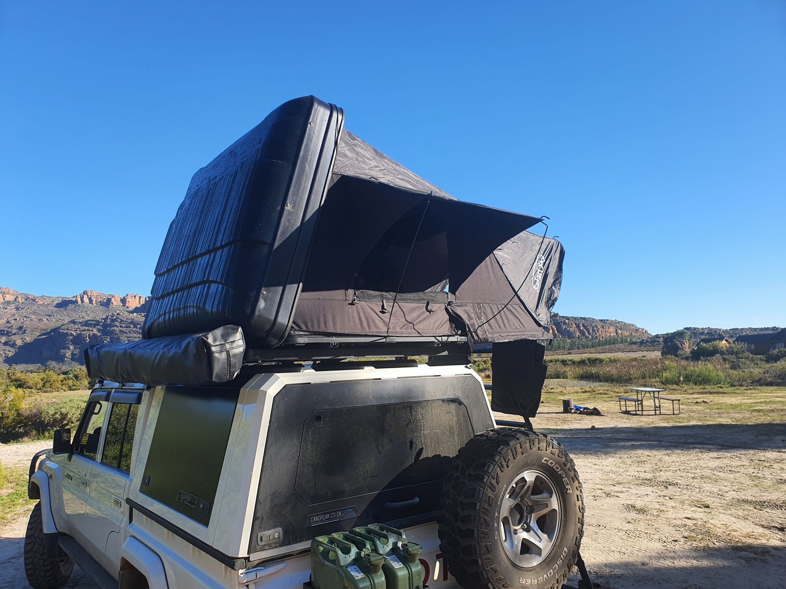 Rooftop tent and canopy open on a white 4x4 with jerry cans below rocky mountains